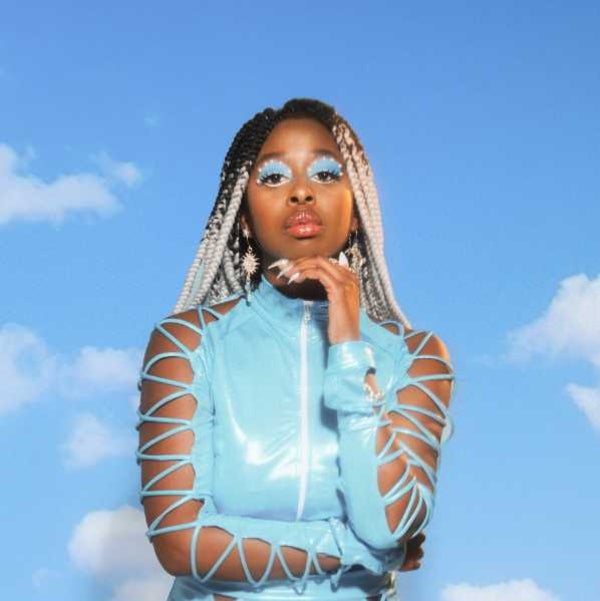 Photo of female musican KYE standing in front of a cloud background wearing a patent blue outfit with matching blue eyeshadow