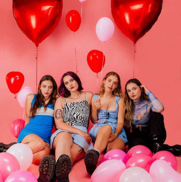 Photo of four female musicians sitting on the ground with pink and red ballons surrounding them