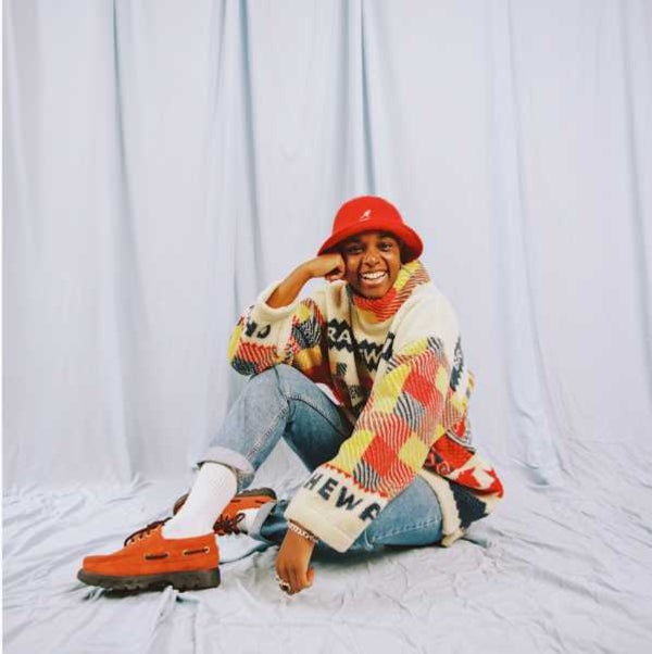 Photo of UK female artist Sherelle sitting on the floor swearing bright coloured street clothes and smiling